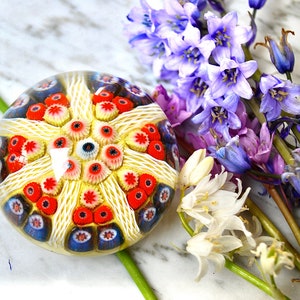 May include: A colorful glass paperweight with a white, red, and blue pattern. The paperweight is sitting on a marble surface with a bouquet of blue and white flowers.