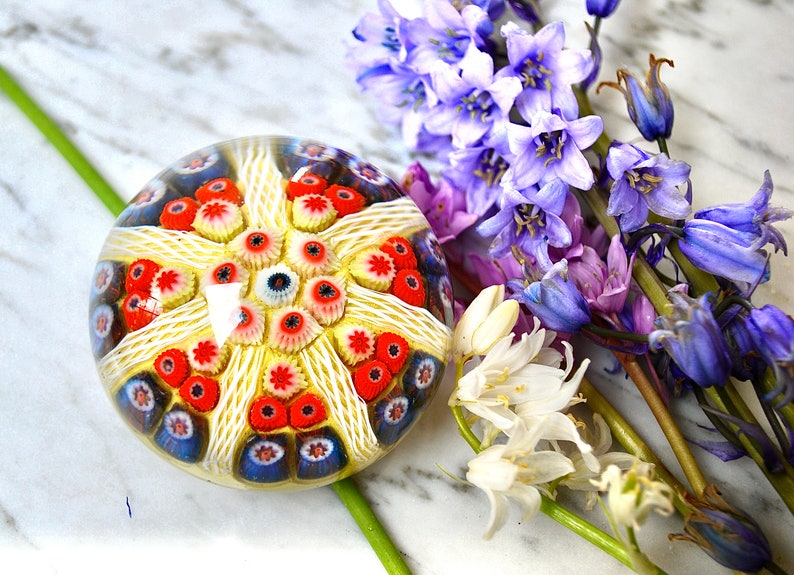 May include: A colorful glass paperweight with a white, red, and blue pattern. The paperweight is sitting on a marble surface with a bouquet of blue and white flowers.