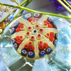 May include: A clear glass paperweight with a colorful, intricate design. The design features a central starburst with red, white, and blue accents.