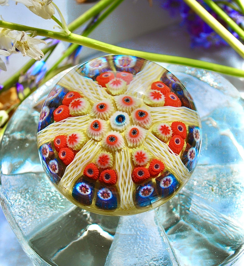 May include: A clear glass paperweight with a colorful, intricate design. The design features a central starburst with red, white, and blue accents.