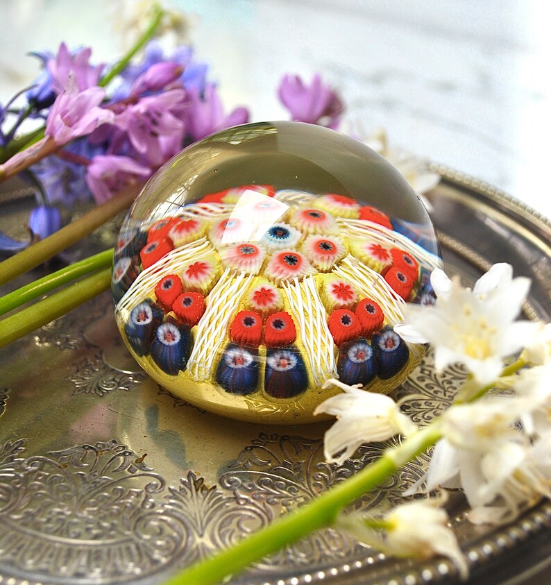 May include: A glass paperweight with a colorful, multi-layered design of red, white, and blue flowers. The paperweight is sitting on a silver tray with a floral design.