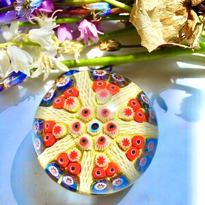 May include: A colorful glass paperweight with a white, yellow, red, and blue floral pattern. The paperweight is round and has a clear glass base.