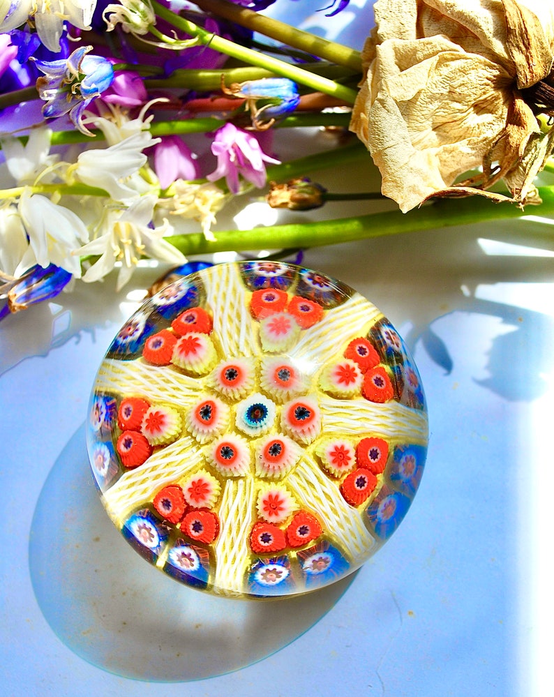 May include: A colorful glass paperweight with a white, yellow, red, and blue floral pattern. The paperweight is round and has a clear glass base.