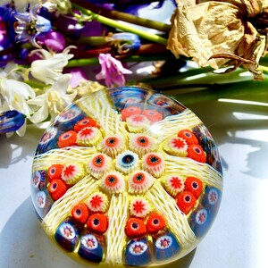 May include: A colorful glass paperweight with a yellow, white, red, and blue design. The paperweight is round and has a textured surface.
