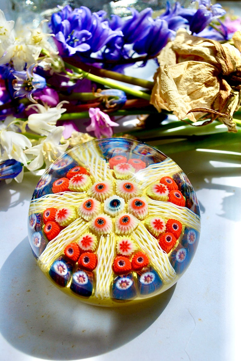 May include: A colorful glass paperweight with a yellow, white, red, and blue design. The paperweight is round and has a textured surface.