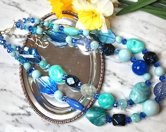Op de afbeelding: Een dubbele ketting met blauwe en groene glazen kralen. De ketting is gemaakt van kralen in verschillende vormen en maten, waaronder ronde, ovale en druppelvormige kralen. De kralen zijn in een willekeurig patroon gerangschikt.
