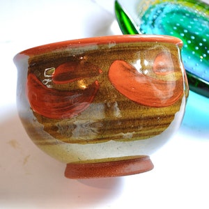 Puede incluir: Un cuenco de cerámica marrón con un esmalte marrón claro y diseños pintados de color naranja. El cuenco tiene una forma redondeada y un borde ligeramente acampanado.
