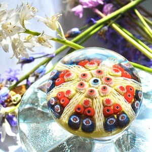 May include: A clear glass paperweight with a colorful, multi-layered design of red, white, and blue. The design features a central starburst with smaller, circular patterns around it.