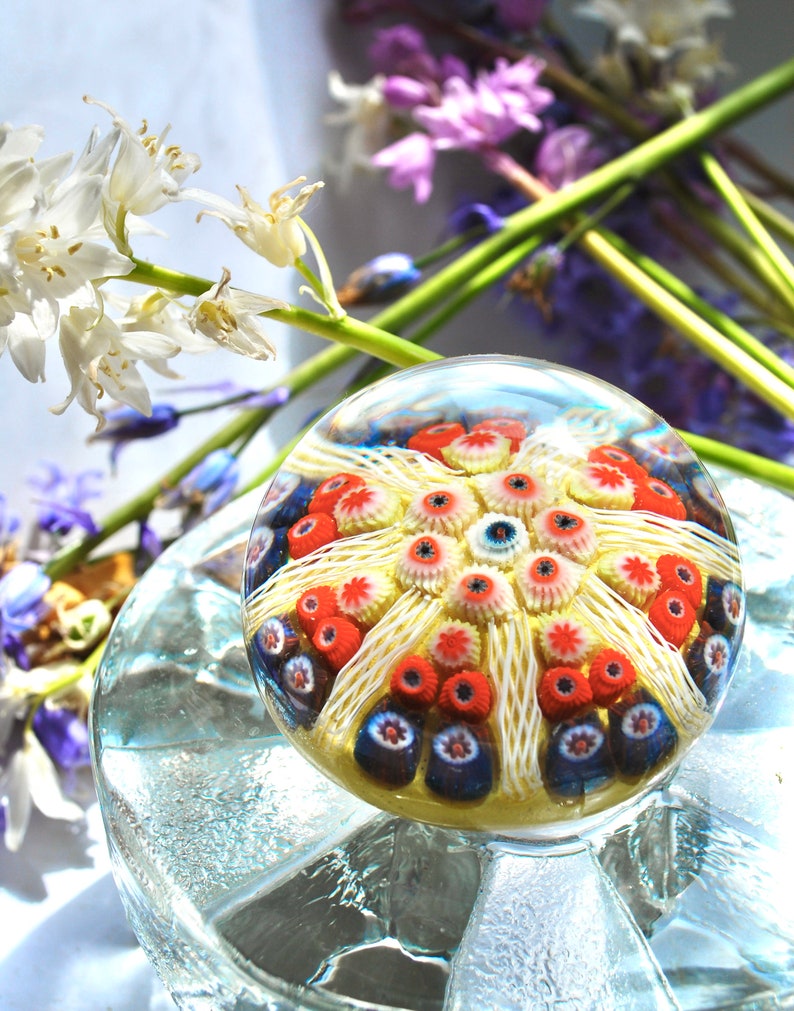 May include: A clear glass paperweight with a colorful, multi-layered design of red, white, and blue. The design features a central starburst with smaller, circular patterns around it.