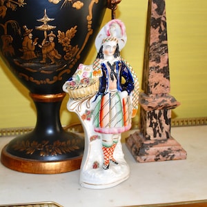 Puede incluir: Una figurita de porcelana de una persona que lleva una chaqueta azul, una falda de cuadros rojos y verdes y un sombrero blanco con una pluma. La figurita sostiene una cesta de flores y está de pie sobre una base blanca.