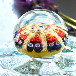 May include: A clear glass paperweight with a colorful, intricate design inside. The design features white, red, and blue glass flowers with a yellow background.