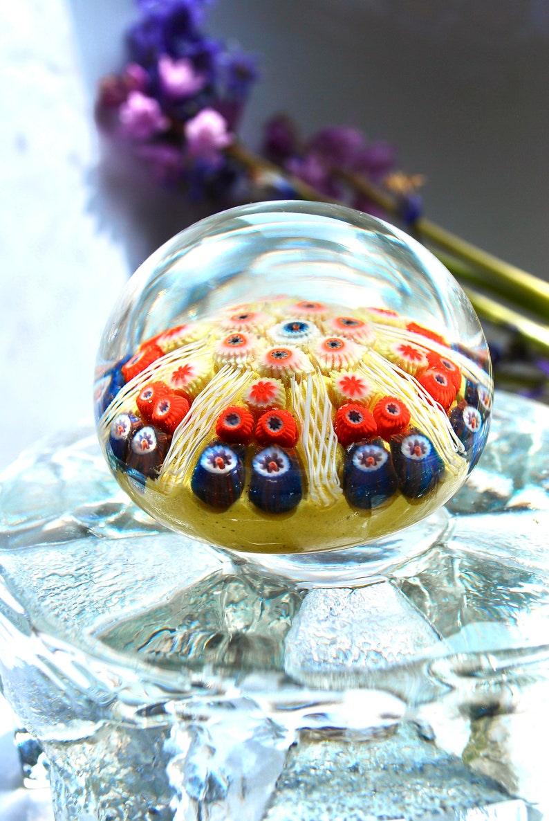 May include: A clear glass paperweight with a colorful, intricate design inside. The design features white, red, and blue glass flowers with a yellow background.