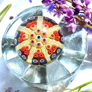 May include: A colorful glass paperweight with a white, red, and blue starburst design. The paperweight is sitting on a clear glass base.