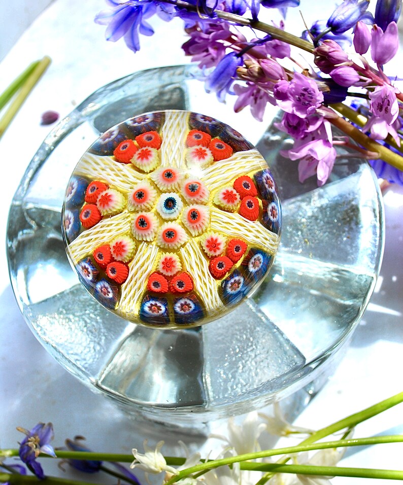 May include: A colorful glass paperweight with a white, red, and blue starburst design. The paperweight is sitting on a clear glass base.