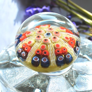 May include: A clear glass paperweight with a colorful, multi-layered design. The design features a central yellow flower with white petals and red and blue accents.