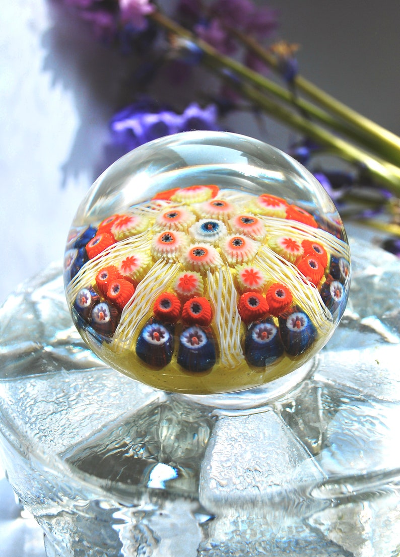May include: A clear glass paperweight with a colorful, multi-layered design. The design features a central yellow flower with white petals and red and blue accents.