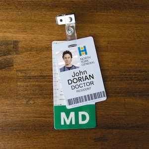 Peut inclure: Un badge d'identification en plastique blanc avec une photo d'un homme portant une blouse bleue. Le badge indique "North York General" et "John Dorian Doctor Resident". Le badge est suspendu à une sangle en plastique transparente avec un clip métallique. Le badge est sur une surface en bois brun.