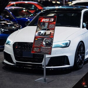 May include: A white Audi RS 3 car is displayed with a sign. The sign has the text "RS 3" in red and white. The sign also has information about the car's stats and origin. The car is parked in a showroom.