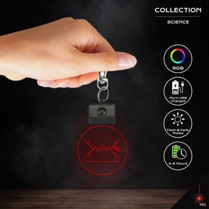 May include: A black science-themed keyring with a red illuminated diagram. The keyring has a silver clasp and a black rectangular light housing. The image includes the text "COLLECTION SCIENCE", and icons for RGB, Micro USB charging, flash & fade modes, and 4-6 hours of use.