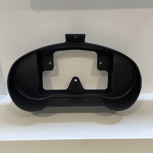 May include: Black plastic automotive gauge pod with three mounting holes. The pod is designed to hold a gauge cluster.