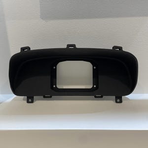 May include: Black plastic automotive gauge bezel with a rectangular cutout for a gauge. The bezel has a curved top and multiple mounting tabs.