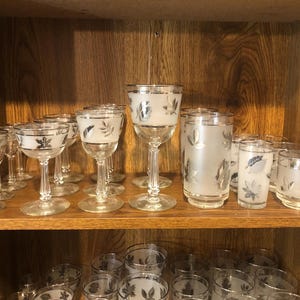 May include: A collection of vintage glassware with a leaf design. The set includes various sizes of stemmed glasses, tumblers, and shot glasses, all with silver rims and frosted leaf patterns. The glasses are displayed on a wooden shelf.
