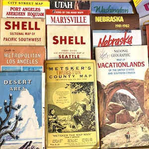May include: A collection of vintage paper road maps including titles such as Shell Sectional Map of Pacific Southwest, Shell Street Guide of Seattle, Metsker's Thurston County Map, and a National Geographic Map of Vacationlands featuring a desert arch.