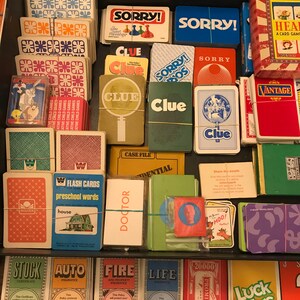 May include: A close-up view of a collection of vintage board games and card games. The games include Clue, Sorry!,  and a game called "Heal". The games are arranged in a drawer and are in various colors, including red, blue, green, and yellow.