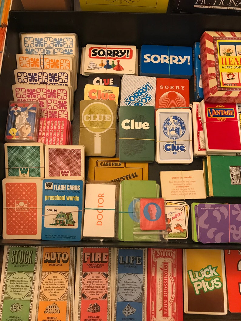 May include: A close-up view of a collection of vintage board games and card games. The games include Clue, Sorry!,  and a game called "Heal". The games are arranged in a drawer and are in various colors, including red, blue, green, and yellow.