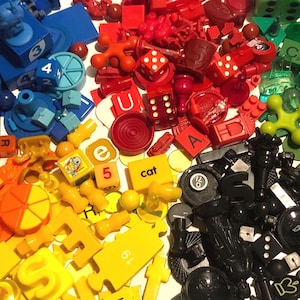 May include: A collection of assorted game pieces and tokens in a vibrant spectrum of colors including blue, red, green, yellow, orange, and black. The assortment features dice, letter tiles, puzzle pieces, and various plastic shapes scattered together.