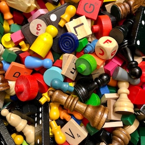 May include: A collection of assorted game pieces including black dominoes with white dots, wooden chess pieces in natural and dark brown, colorful plastic pawns, and square letter tiles with printed characters like G, L, D, B, and V.