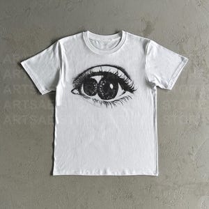 May include: White t-shirt with a black and white illustration of a single eye with two pupils.