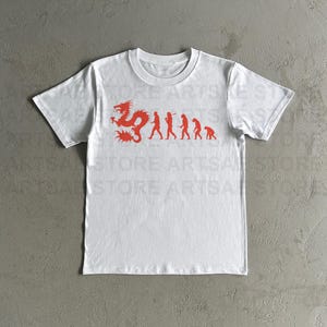 Peut inclure: T-shirt blanc avec un dragon rouge et une ligne de silhouettes rouges marchant en ligne. Les figures sont à différents stades d'évolution, d'un singe primitif à un humain moderne.