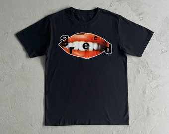Op de afbeelding: Zwart T-shirt met een afbeelding van een rode mond met witte tanden en het woord "greed" gedrukt in zwarte letters.