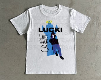 Op de afbeelding: Wit T-shirt met een blauwe en paarse illustratie van een persoon die op een stoel zit met een halo boven zijn hoofd. De tekst "LUCKI" is in zwart boven de illustratie gedrukt. De tekst "FREE WAVE 3" is in zwart onder de illustratie gedrukt.