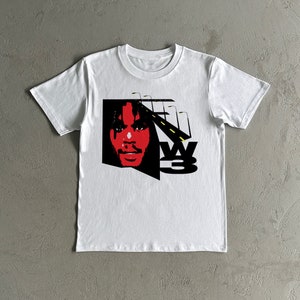 以下が含まれることがあります： 白地に黒と赤のグラフィックプリントが施されたTシャツ。顔と「W3」の文字がプリントされている。