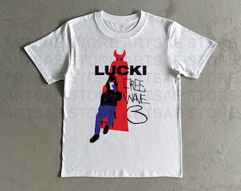 Op de afbeelding: Wit T-shirt met een zwarte en rode grafische print van een persoon die op een stoel zit met een rode duivel erachter. De tekst "LUCKI FREE WAVE 3" is op het T-shirt gedrukt.