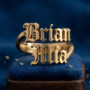 May include: A gold-toned metal ring featuring two stacked names, Brian and Mia, written in a stylized gothic font. The ring has a polished finish and is designed with an open band structure, displaying the names clearly across the front surface.