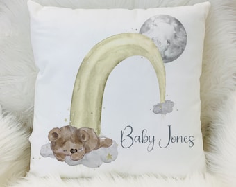 May include: A square white throw pillow featuring a watercolor illustration of a brown teddy bear sleeping on a gray cloud. A pale yellow rainbow arches over the bear toward a gray moon. The text Baby Jones is printed in dark gray script.