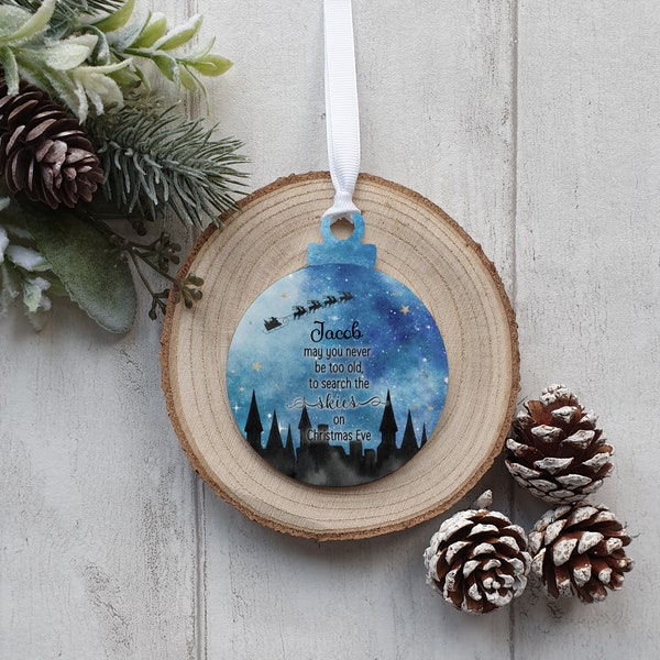 May include: A blue Christmas ornament with a white ribbon, featuring a night sky design with Santa's sleigh and text. The ornament is placed on a wooden slice, with pine cones and greenery in the background.