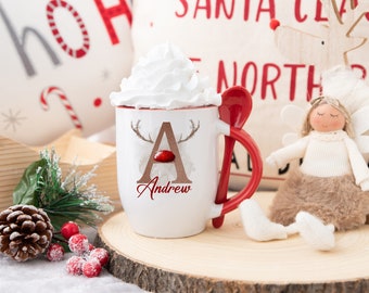 May include: A white ceramic mug with a red interior, filled with whipped cream, and a red spoon attached to the handle. The mug features a brown letter "A" with reindeer antlers and the name "Andrew" in red. Christmas-themed decor surrounds the mug.