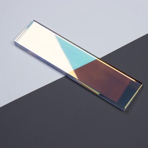 Op de afbeelding: Een lange, rechthoekige prisma van iriserend glas met een kleurverloop, waaronder blauw, groen en paars. De glazen prisma ligt op een zwart oppervlak.