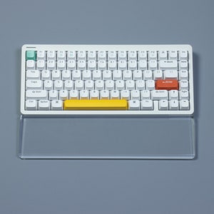 May include: A white mechanical keyboard with a yellow space bar and an orange enter key. The keyboard has a clear acrylic wrist rest.