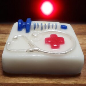May include: White soap bar with a red cross, a silver stethoscope, and a blue syringe on top. The soap bar is on a wooden surface.