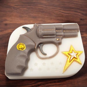 May include: A gray revolver-shaped soap with gold accents and a gold star on a white base. The soap has a detailed design and is set against a dark wood background.