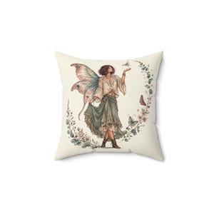 May include: A square throw pillow featuring a cream-colored background with a central illustration of a figure with large, detailed moth-like wings, wearing a light-colored blouse and a long, layered green skirt, surrounded by a floral wreath.