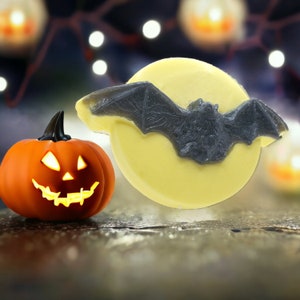 May include: A yellow and white Halloween-themed soap bar with a black bat design. The soap bar is next to a small orange pumpkin with a carved jack-o'-lantern face.