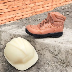 May include: A brown work boot and a white hard hat on a concrete surface. The boot is made of leather and has a black sole. The hard hat is made of plastic and has a white strap.