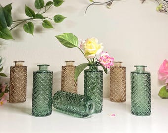 May include: A collection of decorative glass vases in shades of green and brown, arranged on a white surface. One vase holds a small bouquet of yellow and pink flowers. The vases have a diamond pattern.