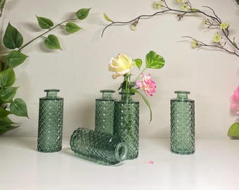 May include: A collection of five green glass bud vases with a diamond pattern. One vase is lying on its side. Two vases hold flowers with yellow and pink blooms. Decorative floral arrangement.
